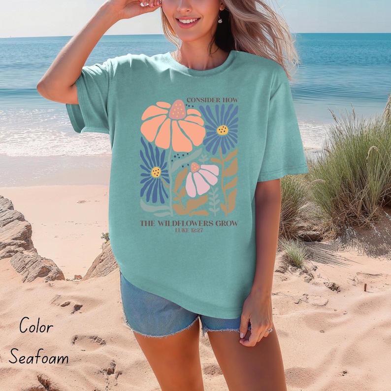 K&ouml;nnte beinhalten: T-Shirt in Seetanggr&uuml;n mit einem Blumenmuster und dem Text "Betrachte, wie die Wildblumen wachsen Lukas 12:27".