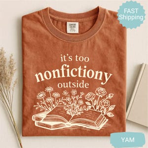 Op de afbeelding: Een roestkleurig t-shirt met de witte tekst "it's too nonfictiony outside" en een bloemenillustratie van een open boek. Het shirt heeft een ronde hals en korte mouwen. Het Comfort Colors-label is zichtbaar.