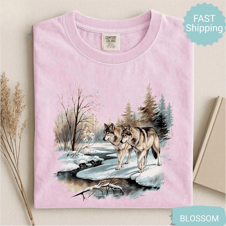 Op de afbeelding: Lichtroze T-shirt met een winterse sc&egrave;ne van twee wolven bij een besneeuwde rivier. De afbeelding bevat bomen en een besneeuwd landschap. Het shirt heeft een Comfort Colors label.