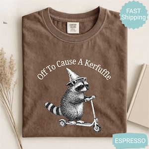 May include: Espresso-colored t-shirt with the phrase "Off To Cause A Kerfuffle" in a curved white font. The graphic features a raccoon wearing a wizard hat and riding a scooter. The shirt is made of Comfort Colors material.