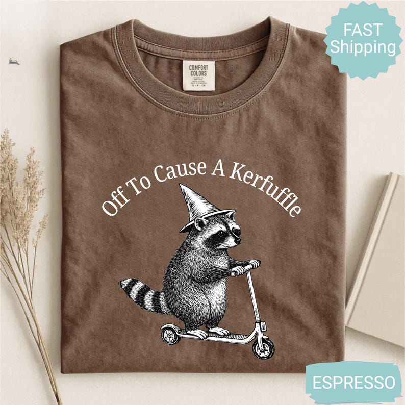 May include: Espresso-colored t-shirt with the phrase "Off To Cause A Kerfuffle" in a curved white font. The graphic features a raccoon wearing a wizard hat and riding a scooter. The shirt is made of Comfort Colors material.