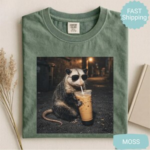May include: Moss green t-shirt featuring a graphic of an opossum wearing sunglasses and drinking an iced coffee. The opossum is set against a blurred urban background. The shirt has a Comfort Colors tag.