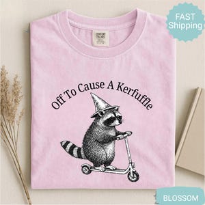May include: Light pink t-shirt with the phrase "Off To Cause A Kerfuffle" above a raccoon wearing a witch hat and riding a scooter. The raccoon illustration is in black and white. The shirt has a crew neck and short sleeves.