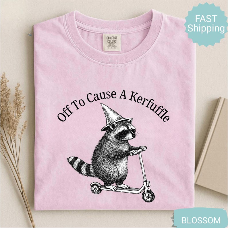 May include: Light pink t-shirt with the phrase "Off To Cause A Kerfuffle" above a raccoon wearing a witch hat and riding a scooter. The raccoon illustration is in black and white. The shirt has a crew neck and short sleeves.