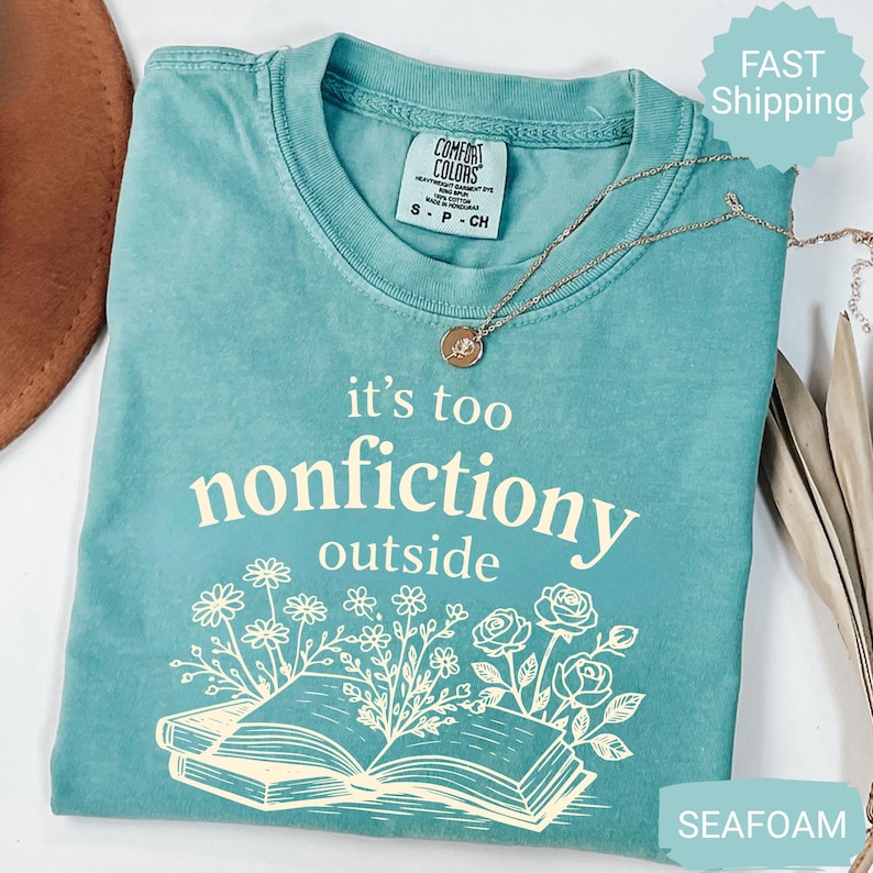 Op de afbeelding: Zeegroen t-shirt met de witte tekst "it's too nonfictiony outside". Het ontwerp bevat een open boek met bloemenillustraties. Een gouden ketting met een hanger ligt op het shirt.