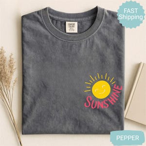 May include: A grey t-shirt featuring a yellow smiling sun graphic and the word "Sunshine" in pink. The shirt has a crew neck and a small tag at the neckline. The shirt is folded and has a "Fast Shipping" sticker.