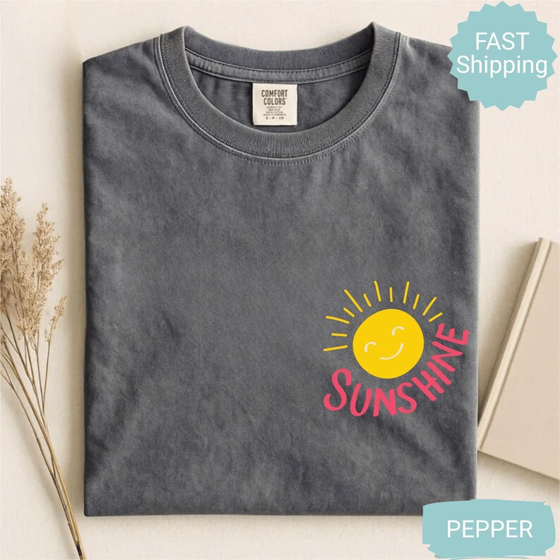 May include: A grey t-shirt featuring a yellow smiling sun graphic and the word "Sunshine" in pink. The shirt has a crew neck and a small tag at the neckline. The shirt is folded and has a "Fast Shipping" sticker.