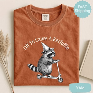 Könnte beinhalten: Rostfarbenes T-Shirt mit dem Schriftzug "Off To Cause A Kerfuffle" über einem Waschbären auf einem Roller, der einen Zaubererhut trägt. Die Grafik ist in Schwarz-Weiß gehalten. Das Shirt hat einen Rundhalsausschnitt.