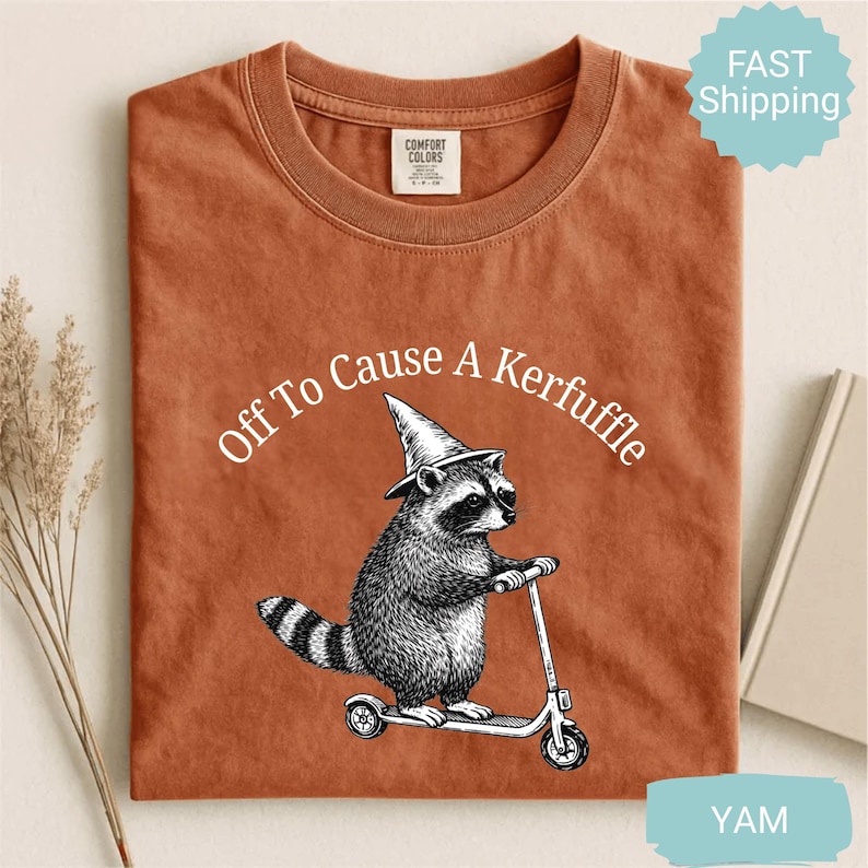 May include: A rust-colored t-shirt with the phrase "Off To Cause A Kerfuffle" arched above a raccoon riding a scooter and wearing a wizard hat. The graphic is in black and white. The shirt has a crew neck.