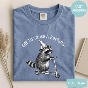 May include: Blue jean colored t-shirt with a graphic of a raccoon wearing a wizard hat and riding a scooter. The text "Off To Cause A Kerfuffle" is arched above the raccoon. The shirt has a crew neck and short sleeves.