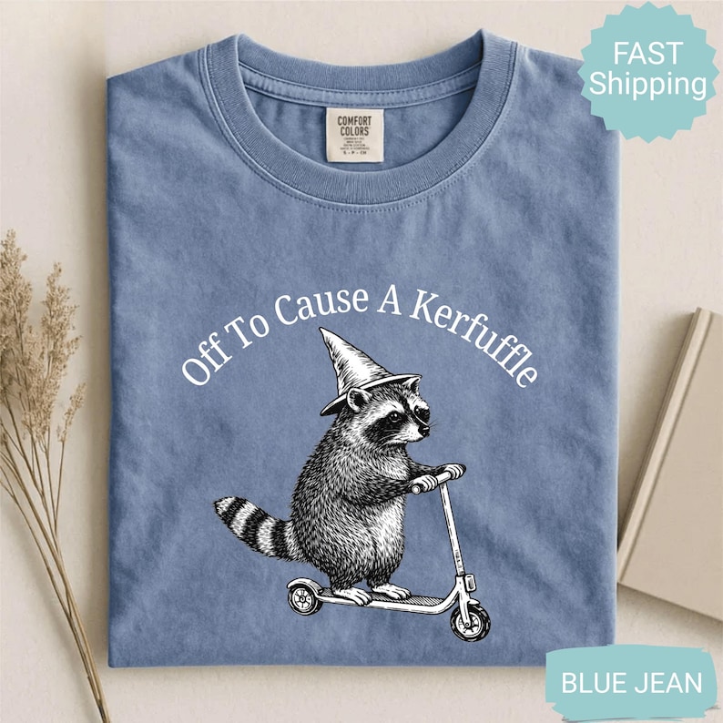 May include: Blue jean colored t-shirt with a graphic of a raccoon wearing a wizard hat and riding a scooter. The text "Off To Cause A Kerfuffle" is arched above the raccoon. The shirt has a crew neck and short sleeves.