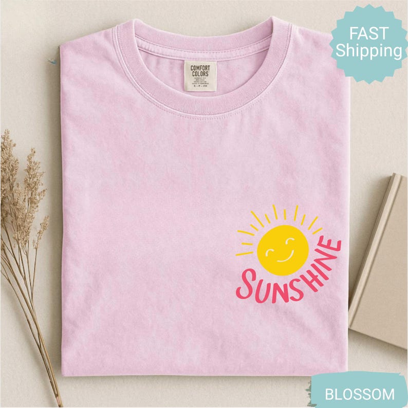 May include: A pale pink t-shirt with a crew neck and short sleeves. The shirt features a yellow smiling sun graphic with the word "SUNSHINE" in pink. The shirt is made of a soft material and has a "Comfort Colors" tag.