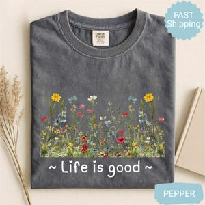 May include: A grey Comfort Colors t-shirt with a floral design featuring yellow, blue, red, and white flowers. The text "~ Life is good ~" is printed below the flowers. The shirt has a crew neck and short sleeves.