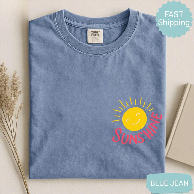 May include: Blue jean coloured t-shirt featuring a yellow smiling sun graphic and the word "SUNSHINE" in pink. The shirt is made of a soft material and has a crew neck. The shirt is folded neatly.