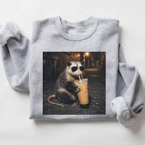 May include: A light gray sweatshirt featuring a graphic of an opossum wearing sunglasses and drinking an iced coffee. The opossum is in an alleyway setting. The sweatshirt is a crew neck style.