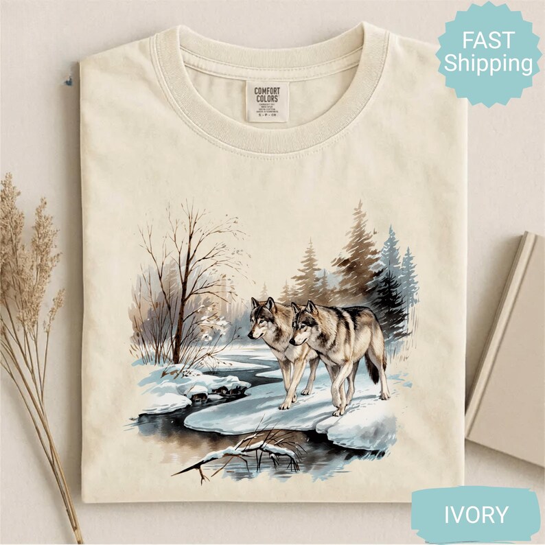 Op de afbeelding: Ivoor Comfort Colors T-shirt met een aquarel illustratie van twee wolven in een besneeuwd bos. De wolven staan bij een beek met bomen op de achtergrond. Het shirt heeft een klassieke ronde hals.