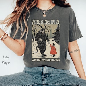May include: Pepper-colored t-shirt featuring a graphic of a horned figure holding hands with a child in a red dress, set in a snowy winter scene. The text "WALKING IN A WINTER WONDERLAND" is printed above and below the illustration.