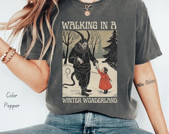 Könnte beinhalten: Dunkelgraues T-Shirt mit einem Grafikdruck, der eine gehörnte Figur zeigt, die die Hand eines Kindes in einem roten Kleid in einer Winterszene hält. Der Text "WALKING IN A WINTER WONDERLAND" steht ober- und unterhalb der Illustration.