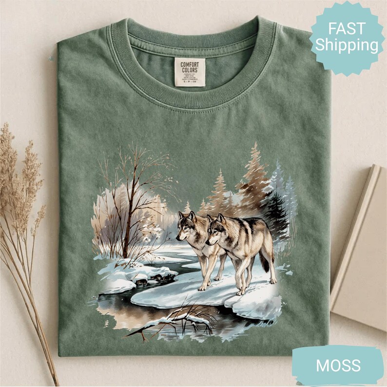 Op de afbeelding: Mosgroen t-shirt met een winterlandschap met twee wolven bij een besneeuwde rivier. Het shirt heeft een ronde hals en een label "Comfort Colors".
