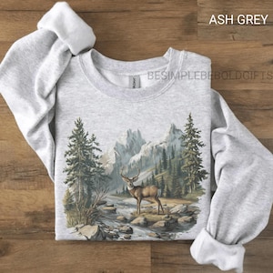 Puede incluir: Sudadera gris ceniza con un paisaje de monta&ntilde;a y bosque, con un ciervo cerca de un r&iacute;o. El texto "BESIMPLEBEBOLDGIFTS" est&aacute; impreso en la sudadera. Tiene mangas largas y cuello redondo.