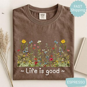 May include: Espresso-coloured t-shirt with a floral design featuring yellow, blue, red, and white flowers. The text "Life is good" is printed in white below the flowers. The shirt has a crew neck and short sleeves.