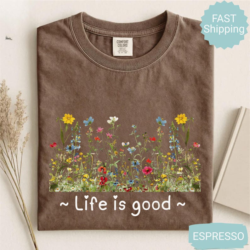 May include: Espresso-coloured t-shirt with a floral design featuring yellow, blue, red, and white flowers. The text "Life is good" is printed in white below the flowers. The shirt has a crew neck and short sleeves.