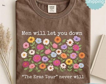 May include: Espresso-colored t-shirt with a floral design and the text "Men will let you down" above "The Eras Tour" never will. The shirt features a crew neck and a Comfort Colors tag.