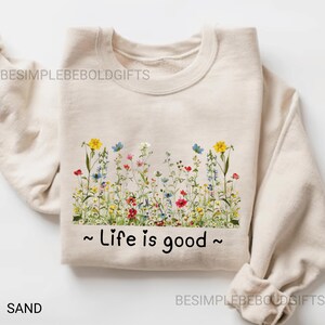 May include: A sand-coloured sweatshirt with a floral design across the chest. The design features yellow, blue, red, and white flowers. The phrase "Life is good" is printed in black below the flowers. The sweatshirt is folded.