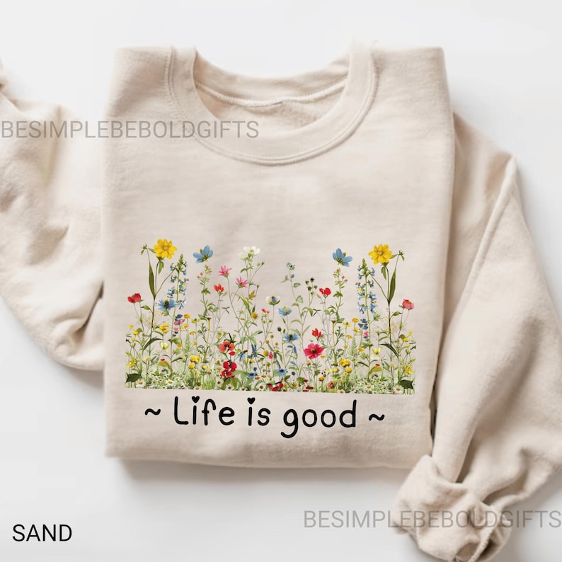 May include: A sand-coloured sweatshirt with a floral design across the chest. The design features yellow, blue, red, and white flowers. The phrase "Life is good" is printed in black below the flowers. The sweatshirt is folded.