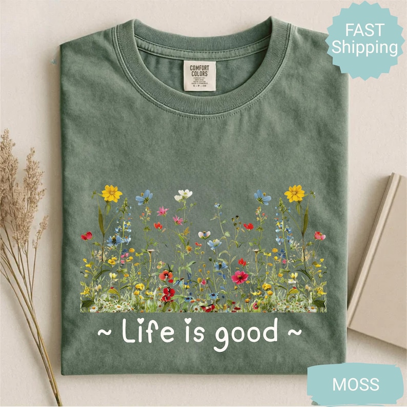 May include: A moss green t-shirt with a floral design. The design includes yellow, blue, red, and white flowers. The text "~ Life is good ~" is printed in white. The shirt has a crew neck and short sleeves.