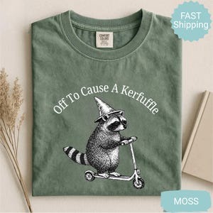 May include: Moss green t-shirt with a graphic of a raccoon wearing a wizard hat and riding a scooter. The text "Off To Cause A Kerfuffle" is arched above the raccoon. The shirt has a crew neck and short sleeves.