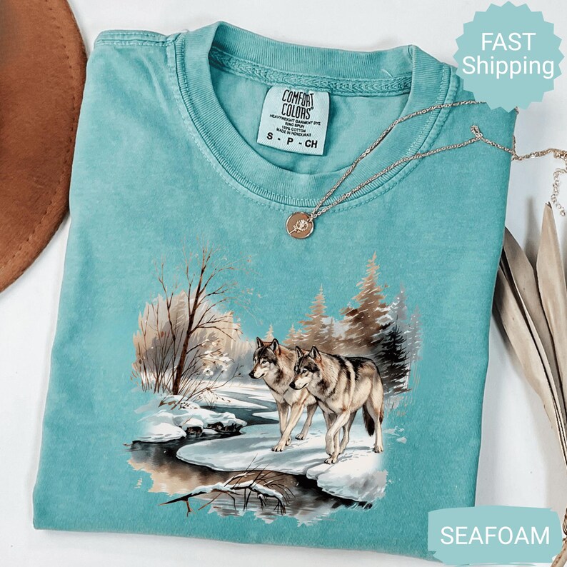 Op de afbeelding: Zeegroen Comfort Colors T-shirt met een winterwolfsc&egrave;ne. De afbeelding toont een besneeuwd landschap met bomen en een rivier. Een zilveren ketting met een hanger ligt op het shirt.