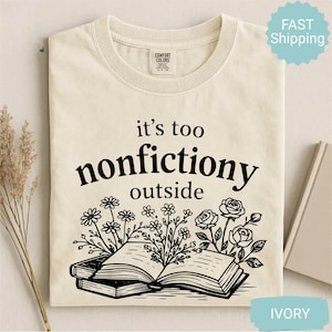 Op de afbeelding: Ivoor kleurig t-shirt met de zwarte tekst "it's too nonfictiony outside". Het ontwerp bevat een open boek met bloemen illustraties. Het t-shirt is opgevouwen.