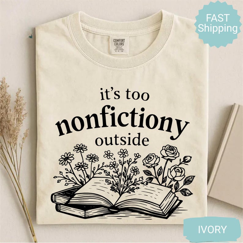 Op de afbeelding: Ivoor kleurig t-shirt met de zwarte tekst "it's too nonfictiony outside". Het ontwerp bevat een open boek met bloemen illustraties. Het t-shirt is opgevouwen.