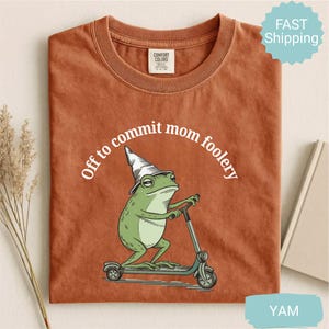 May include: Rust-colored t-shirt featuring a green frog wearing a wizard hat and riding a scooter. The text "Off to commit mom foolery" is arched above the frog. The shirt has a Comfort Colors tag.