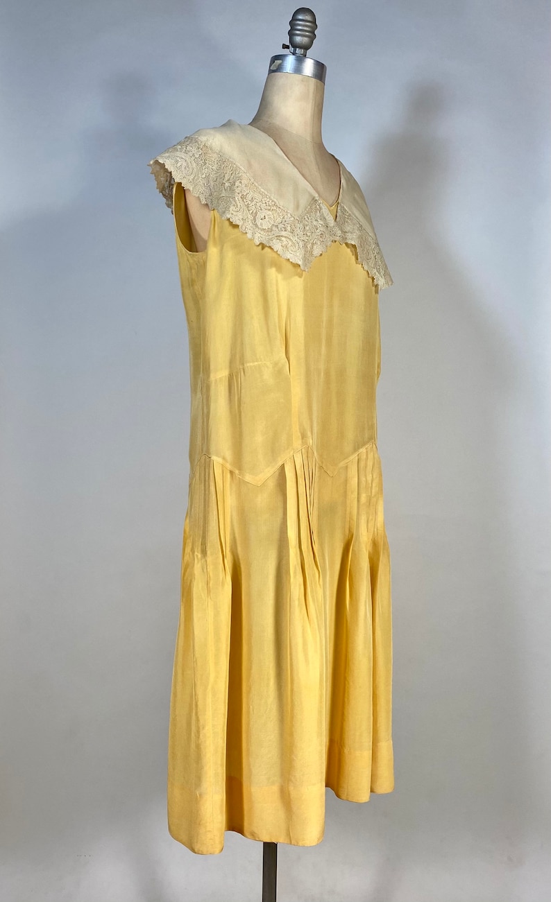 May include: A sleeveless yellow silk dress with a white lace collar. The dress has a V-neckline and a pleated skirt.