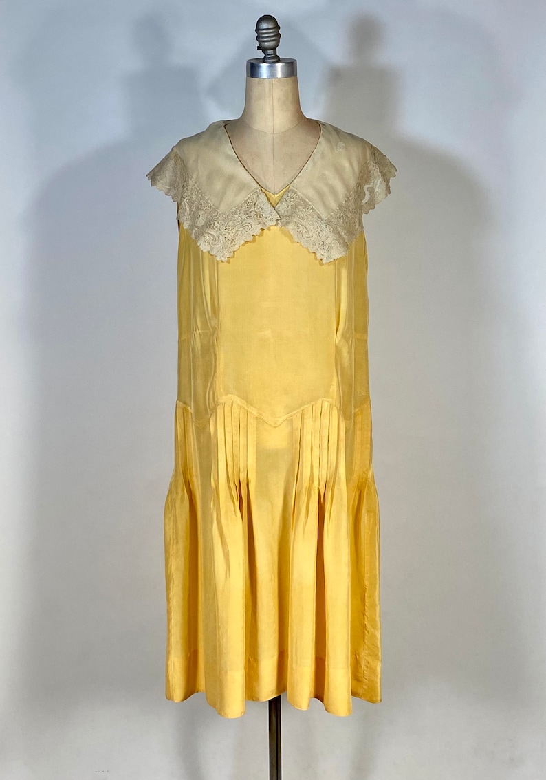 May include: A sleeveless yellow dress with a white lace collar. The dress has a pleated skirt and a fitted bodice.