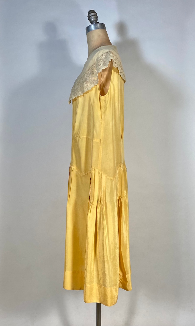 May include: A yellow sleeveless dress with a white lace collar. The dress has a pleated skirt and is made of a silky fabric.