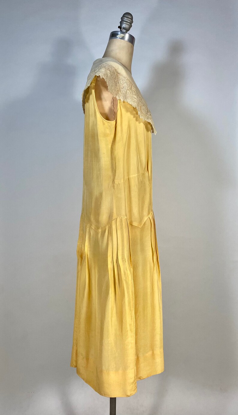 May include: A yellow sleeveless dress with a lace collar. The dress has a pleated skirt and is on a mannequin.