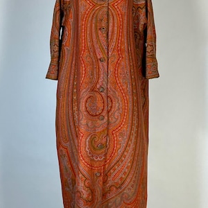 May include: A long, orange paisley patterned coat with a button-up front and long sleeves. The coat is displayed on a mannequin.