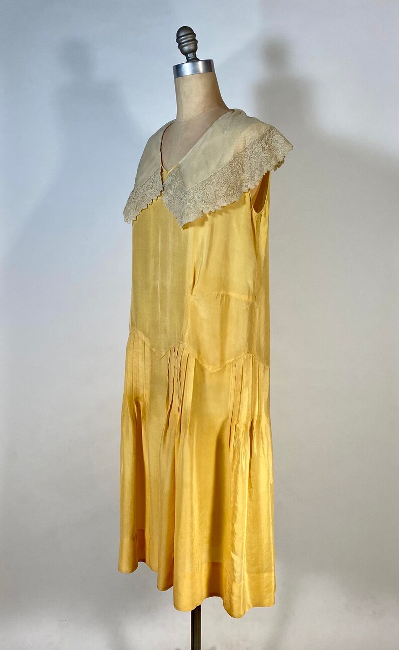 May include: A yellow silk dress with a white lace collar. The dress has a pleated skirt and a sleeveless design.