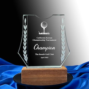 Puede incluir: Un trofeo de vidrio transparente con una base de madera. El trofeo presenta una bola de golf y un palo de golf con un diseño de hojas en los lados. El texto en el trofeo dice "California Korean Championship Tournament" y "Champion". El texto en la parte inferior dice "The Bendit Golf Club" y "Abril 2023".