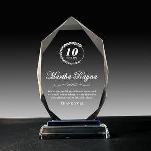 Puede incluir: Un premio de cristal transparente con un diseño facetado. El premio tiene una base azul y presenta el texto "10 YEARS" y "Martha Rayna". El texto "THANK YOU!" también es visible.