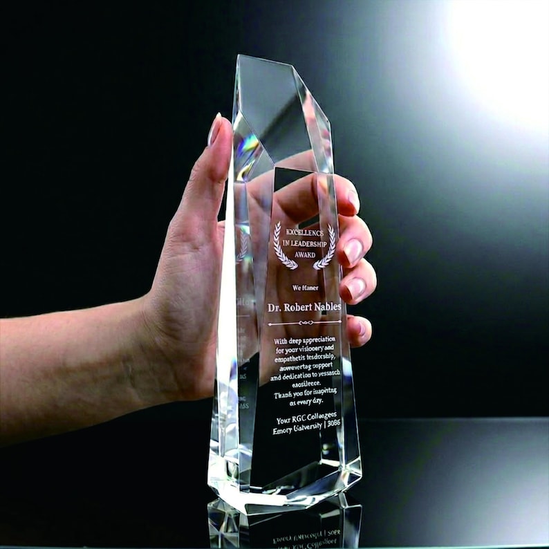 May include: A clear, faceted glass award trophy with a pointed top. The front surface is engraved with the text Excellence in Leadership Award, Dr. Robert Nables, and a message of appreciation from RGC Colleagues at Emory University.