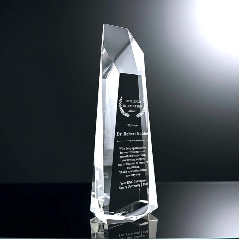 May include: A clear, faceted crystal award trophy with a tall, tapered, geometric shape. A black panel on the front features white engraved text reading Excellence in Leadership Award, Dr. Robert Nables, and a message of appreciation from RGC colleagues.