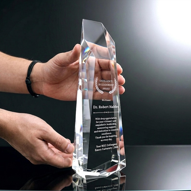 May include: A clear, faceted crystal award trophy with a tapered, geometric shape. The front surface features etched text reading Excellence in Leadership Award, We Honor Dr. Robert Nables, and a message of appreciation from RGC Colleagues.