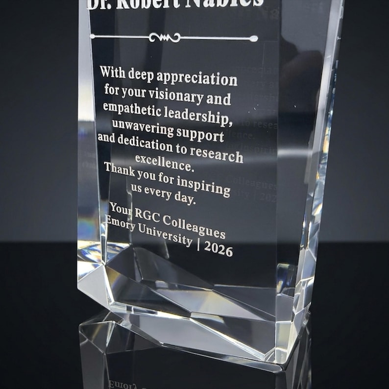 May include: A clear, faceted glass award trophy featuring an engraved inscription that reads: Dr. Robert Nables, With deep appreciation for your visionary and empathetic leadership, unwavering support and dedication to research excellence. Thank you for inspiring us every day. Your RGC Colleagues Emory University | 2026.