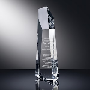 May include: A tall, clear crystal award with a multifaceted, geometric design. The front surface features etched text and a laurel wreath graphic. The award has sharp, angular edges and a reflective, transparent appearance with a pointed top.