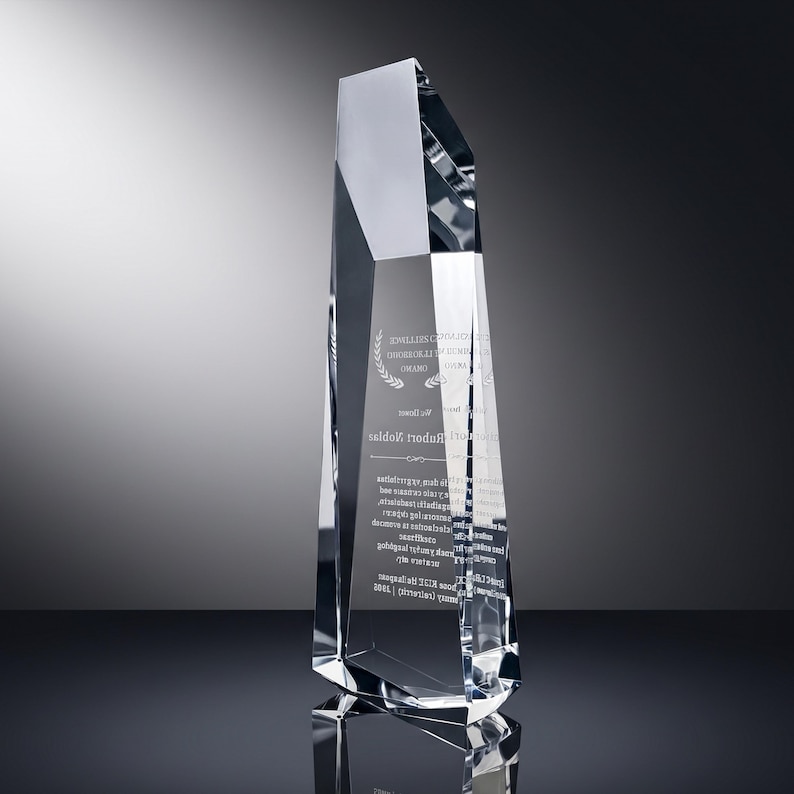 May include: A tall, clear crystal award with a multifaceted, geometric design. The front surface features etched text and a laurel wreath graphic. The award has sharp, angular edges and a reflective, transparent appearance with a pointed top.