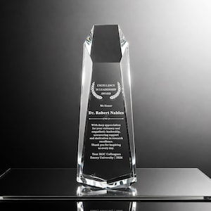 May include: A clear, faceted crystal award trophy with a tapered, multi-sided design. The front surface is engraved with the text: Excellence in Leadership Award, We Honor Dr. Robert Nables, and a message of appreciation from RGC Colleagues.